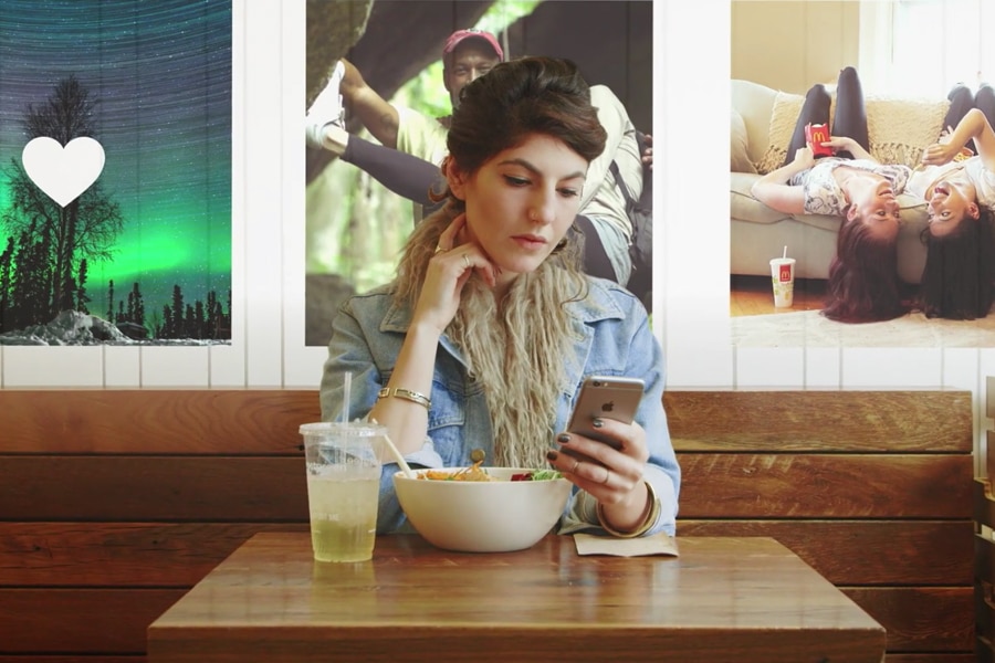 Girl sitting at a table with a bowl of food and a drink, looking at her phone. In the background are photos from Instagram.