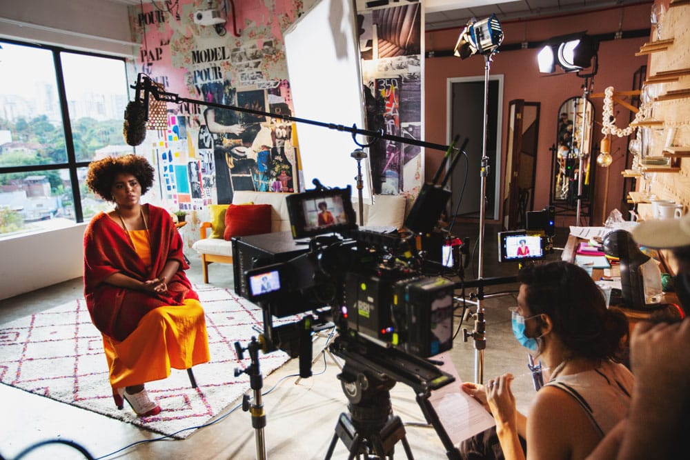 Woman in an orange dress and red jacket sitting down and being filmed with large video equipment in the background.