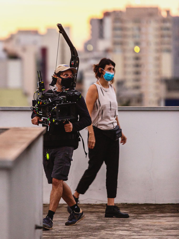 Two people, one holding a large video camera, walking on a rooftop with buildings in the background.